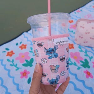 May include: A pink drink sleeve featuring a plaid pattern and cartoon images of Stitch, flowers, and bees. The sleeve is wrapped around a clear plastic cup with a pink straw. The text "shopheavenme" is printed on the sleeve.