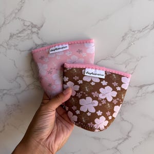 Könnte beinhalten: Zwei Tassenmanschetten aus Neopren mit rosa Rand und weißen Etiketten mit der Aufschrift shophavenrose. Eine Manschette hat einen hellrosa Hintergrund mit weißem Hibiskus-Blumenmuster, die andere einen braunen Hintergrund mit hellrosa Hibiskus-Muster.