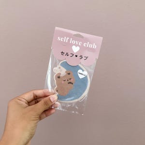 May include: A vanilla-scented air freshener in a clear plastic package. The air freshener is shaped like a bear with a sad expression, on a blue background. The packaging reads "self love club" in pink and white text, with Japanese characters.