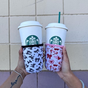 Könnte beinhalten: Zwei weiße To-Go-Becher mit schwarzen Deckeln und grünen Starbucks-Logos. Die Becher werden von einer Person gehalten und haben bunte Stoffhüllen. Eine Hülle hat ein schwarz-weißes Schleifenmuster, die andere ein rosa-rotes Muster mit Herzen und Schleifen.