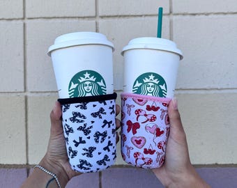 May include: Two white to-go cups with black lids and green Starbucks logos. The cups are in a person's hands and have colorful fabric sleeves. One sleeve has a black and white bow pattern, and the other has a pink and red pattern with hearts and bows.