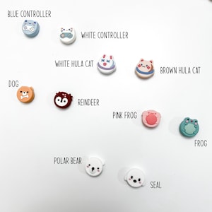 Op de afbeelding: Assortiment van kleurrijke siliconen telefoonbeugels met verschillende dier- en controllerontwerpen. De ontwerpen omvatten een blauwe controller, een witte controller, een witte hula-kat, een bruine hula-kat, een hond, een rendier, een roze kikker, een kikker, een ijsbeer en een zeehond.