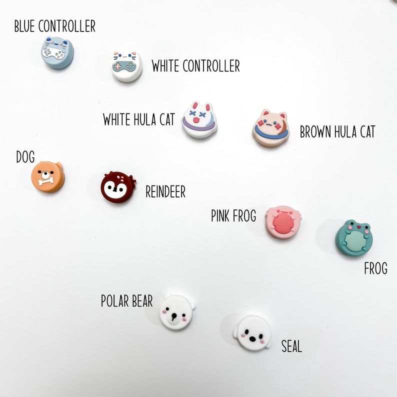 Op de afbeelding: Assortiment van kleurrijke siliconen telefoonbeugels met verschillende dier- en controllerontwerpen. De ontwerpen omvatten een blauwe controller, een witte controller, een witte hula-kat, een bruine hula-kat, een hond, een rendier, een roze kikker, een kikker, een ijsbeer en een zeehond.