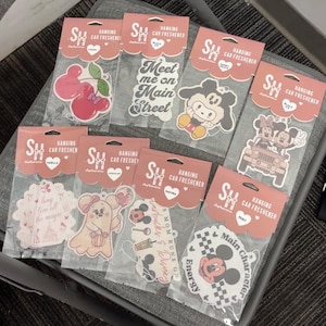 May include: A collection of hanging car air fresheners in various designs. The fresheners feature cartoon characters, cherries, and text like "Meet me on Main Street" and "Main Character". Each is packaged in a clear plastic bag with a pink header.