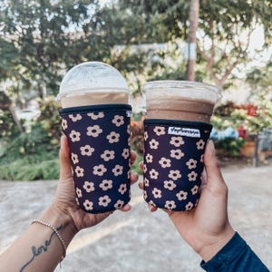 Könnte beinhalten: Zwei Eiskaffeegetränke in transparenten Plastikbechern mit Deckeln, gehalten in marineblauen Blumenprint-Hüllen. Die Hüllen haben eine schwarze Einfassung und ein sich wiederholendes Muster aus beigefarbenen Blumen. Der Hintergrund ist verschwommen mit grünem Laub.