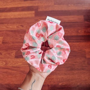 May include: Pink scrunchie with a tropical leaf and flower print. The scrunchie has a small white tag that says "shophawaiiantime".