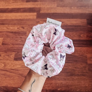 May include: Pink and white scrunchie with a repeating pattern of Hello Kitty and Badtz-Maru characters. The scrunchie has a small white tag with the text "shopheavenmore" on it.