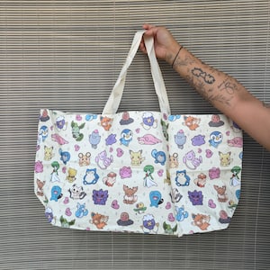 Peut inclure: Un sac fourre-tout blanc orné d'un motif répétitif représentant divers personnages de dessins animés colorés et de petits cœurs roses. Le sac possède deux anses en tissu blanc et est fabriqué dans une matière légère avec un imprimé graphique.