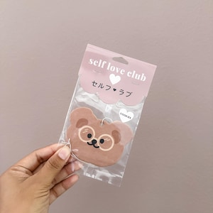 May include: A brown bear-shaped cookie with glasses is packaged in a clear plastic bag. The bag has a pink header with the words "self love club" and Japanese text. The word "COOKIES" is printed on the bag.