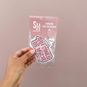 May include: A vanilla-scented hanging car freshener in a clear plastic package. The pink and white freshener has the phrase "Shit Could Be Worse" in a playful font. The packaging is light pink with the words "Hanging Car Freshener".