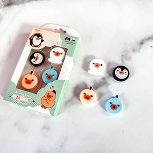 以下が含まれることがあります： ゲームコントローラー用のカラフルなアナログスティックキャップ8個セット。キャップには、ペンギン、タコ、ヒヨコなど、さまざまなかわいい動物のデザインが施されています。パッケージは白地に緑色のアクセントが施され、「Pupu Aliens」と「Analog Caps」の文字が書かれています。