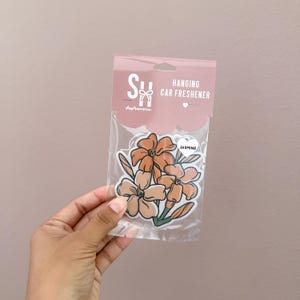 May include: A package of hanging car fresheners featuring a floral design. The fresheners are in a clear plastic bag with a pink header that reads "Hanging Car Freshener." The floral design includes orange and cream-colored flowers with green stems and leaves. The scent is jasmine.
