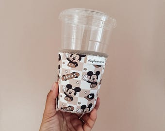 Könnte beinhalten: Beige und weiß gemusterte Becherhülle mit Mickey Mouse-Illustrationen und dem Wort „BRO“ auf einem Skateboard. Die Hülle ist um einen durchsichtigen Plastikbecher mit Deckel gewickelt. Die Hülle hat eine beige Einfassung und der Shop-Name ist auf der Seite aufgedruckt.