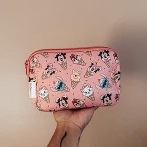May include: Pink zippered cosmetic bag featuring a repeating pattern of cartoon characters on ice cream cones. Characters include Mickey and Minnie Mouse, Stitch, Chip and Dale, Marie, and Winnie the Pooh. The bag has a pink zipper and a small white tag.