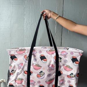 May include: A large tote bag with a light pink background and a repeating pattern of sushi, ramen bowls, and cherry blossoms. The bag has black handles and a black mesh pocket on the side.