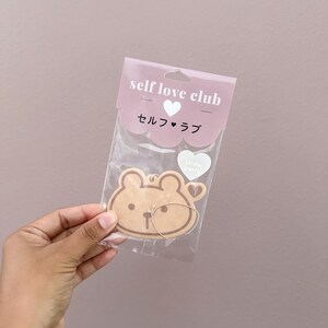 May include: A light brown bear-shaped air freshener with a gold string, packaged in a clear plastic bag. The packaging features the text "self love club" and Japanese characters, along with a heart graphic and the words "strawberry".