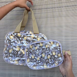 May include: Two clear plastic bags featuring a repeating pattern of cartoon characters and white flowers. The larger bag has two beige fabric handles, while the smaller bag is a rectangular pouch with a beige fabric side tab and a zipper closure.
