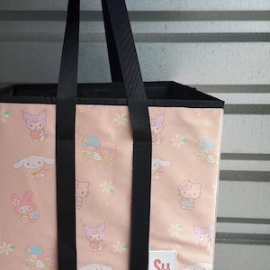 May include: Pink tote bag with black handles featuring a repeating pattern of cartoon characters and flowers. The bag has a rectangular shape and a small white label with the letters "SH".