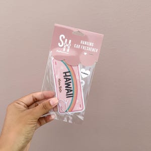 May include: A pink hanging car air freshener with the word "HAWAII" in black capital letters and a rainbow design. The packaging includes the words "Hanging Car Freshener" and "Plumeria".