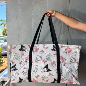May include: White tote bag featuring a repeating pattern of cartoon characters in black, pink, and white. The bag has black handles and a rectangular shape, suitable for carrying various items. The bag is held up by a person's hand.