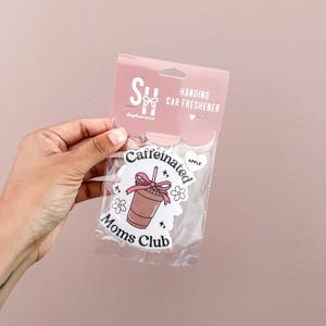 May include: A hanging car air freshener in a clear plastic package. The design features a cartoon coffee cup with a pink bow and the words "Caffeinated Moms Club." The package is pink and has the words "Hanging Car Freshener."