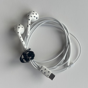 May include: White wired earbuds featuring a black polka dot pattern on the earpieces and the connector housing. The white cable is coiled and secured with a small black plastic clip, keeping the wired audio device neatly organized and compact.