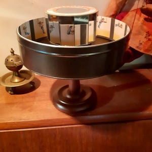 Könnte beinhalten: Ein Vintage-Zoetrop, ein Gerät zur Vorfilm-Animation, mit einer rotierenden Trommel mit sequenziellen Bildern von Figuren in Bewegung. Das Gerät besteht aus Metall und Holz mit dunkelbraunem Finish. Links befindet sich ein kleiner Messing-Räuchergefäß.