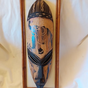 May include: A wooden mask with an elephant carved into it. The mask is painted in shades of brown, black, and blue. It is framed in a wooden frame.