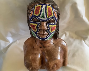 May include: Wooden bust sculpture of a person with a beaded face mask. The mask is made of colorful beads in shades of blue, green, red, yellow, and white. The bust is carved from dark brown wood.
