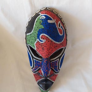 May include: A beaded mask with a black, red, green, and blue design. The mask has a black mouth and eye openings. The mask is decorated with white beads around the edges.