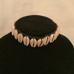 May include: A choker necklace featuring a row of cream-colored cowrie shells. The shells are strung together on a black cord, creating a striking contrast. The necklace is displayed on a black velvet surface.