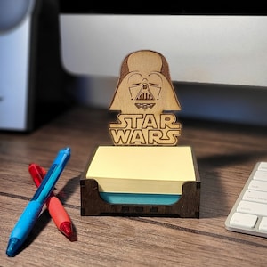 May include: A wooden Star Wars themed note holder with a Darth Vader design. The holder has a yellow sticky note pad and a blue sticky note pad. The holder is on a wooden surface.