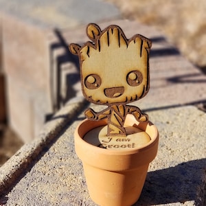 May include: A wooden cutout of a cartoon Groot character from the Guardians of the Galaxy movie series, standing in a small terracotta pot. The pot has the words "I am Groot!" written on it.