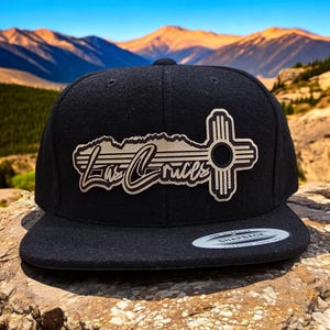May include: Black snapback hat featuring the text "Las Cruces" in a stylized font, along with a graphic of a cross-like symbol. The hat has a flat brim and a classic snap closure.