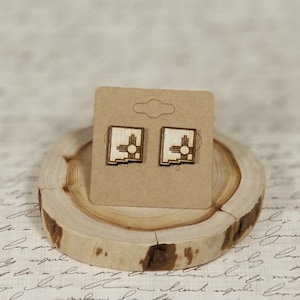 May include: A pair of wooden earrings featuring a laser-cut design of the state of New Mexico with the Zia symbol. The earrings are displayed on a small wooden stand.