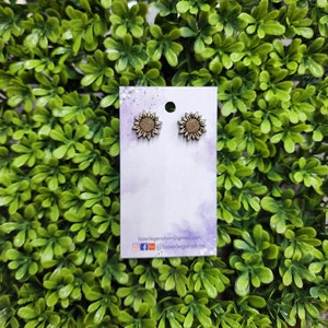 May include: A pair of wooden sunflower earrings on a white card with a purple watercolor background. The earrings are brown and have a light brown center.