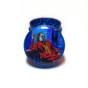 May include: Blue herb grinder with a detailed image of the Joker in a red suit, yellow vest, and green hair. The grinder is made of metal and has a glossy finish. The Joker is seated, holding a cigarette.