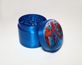 Puede incluir: Un grinder de hierbas azul de tres piezas con una tapa colorida que presenta un diseño abstracto en rojo, naranja y azul. El grinder es de metal y tiene una superficie texturizada.