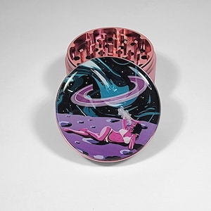 May include: A pink metal herb grinder with a detailed top and a graphic design on the lid. The lid features a woman reclining on a purple surface with a space-themed background.