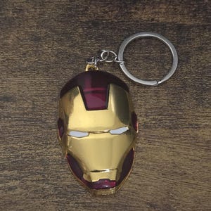 Könnte beinhalten: Ein goldfarbener Iron Man Masken-Schlüsselanhänger mit einem silbernen Schlüsselring. Die Maske hat rote und weiße Akzente. Der Schlüsselanhänger ist ein Sammlerstück.