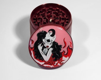 Puede incluir: Un molinillo de metal rojo con una tapa que presenta un gráfico de una mujer con cara de calavera, cabello negro y llamas rojas. El molinillo es redondo con un interior texturizado.