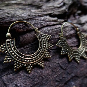 May include: A pair of gold hoop earrings with a detailed, sunburst-like design. The earrings are made of metal and have a circular shape.