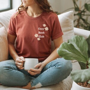 May include: A woman wearing a rust-colored t-shirt with the text "Cozy Book Club" and small white flowers printed on the front. She is sitting on a couch with a white mug in her hands.