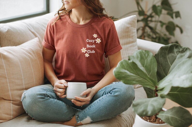 May include: A woman wearing a rust-colored t-shirt with the text "Cozy Book Club" and small white flowers printed on the front. She is sitting on a couch with a white mug in her hands.