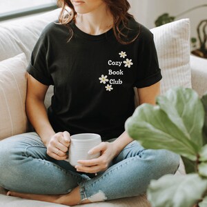 May include: A person wearing a black t-shirt with the text "Cozy Book Club" and white daisy flowers printed on the front. The person is sitting on a couch with a white mug in their hands.