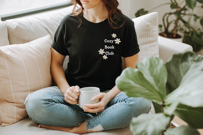 May include: A person wearing a black t-shirt with the text "Cozy Book Club" and white daisy flowers printed on the front. The person is sitting on a couch with a white mug in their hands.