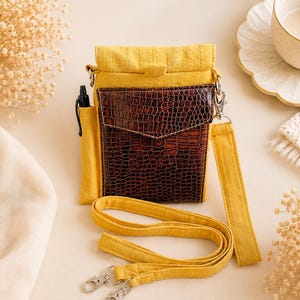 K&ouml;nnte beinhalten: Eine gelbe Stoff-Umh&auml;ngetasche mit einer dunkelbraunen Kunstledertasche auf der Vorderseite, die ein strukturiertes Krokodilledermuster aufweist. Die Tasche hat ein Seitenfach f&uuml;r einen schwarzen Stift und einen gelben Trageriemen mit silbernen Metallclips.