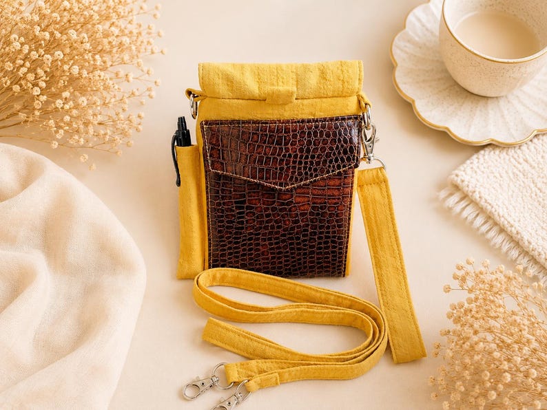 K&ouml;nnte beinhalten: Eine gelbe Stoff-Umh&auml;ngetasche mit einer dunkelbraunen Kunstledertasche auf der Vorderseite, die ein strukturiertes Krokodilledermuster aufweist. Die Tasche hat ein Seitenfach f&uuml;r einen schwarzen Stift und einen gelben Trageriemen mit silbernen Metallclips.