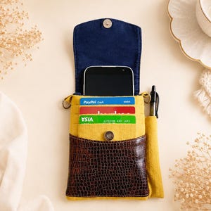 K&ouml;nnte beinhalten: Eine gelbe Handy-Umh&auml;ngetasche mit dunkelblauem Innenklappenteil und einer dunkelbraunen Fronttasche in Krokodillederoptik. Die Tasche enth&auml;lt ein schwarzes Smartphone, drei Kreditkarten und einen schwarzen Stift in einer seitlichen Schlaufe.