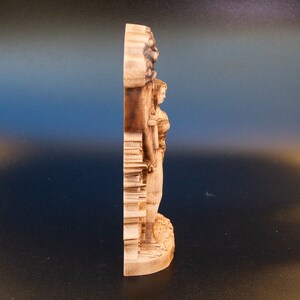 May include: A wooden sculpture of a woman standing with her arms raised, carved in a detailed style. The sculpture is made of light brown wood and has a natural finish.